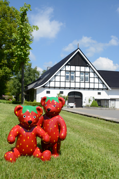 Zwei niedliche ErdBär-Figuren stehen auf dem grünen Rasen. Im Hintergrund ist das Fachwerkhaus des Erdbeerhofes Böckmann zu sehen.