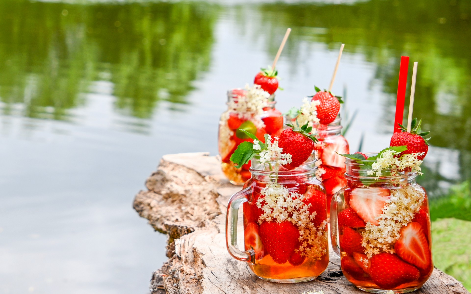 Vier Glas-Henkelbecher gefüllt mit frischen, roten Erdbeeren und Holunderblüte und einem Pfefferminzblatt als Topping zeigen wunderbare Sommer-Cocktails. In den Gläsern steckt ein roter Strohhalm und ein Holzspießer. Die Gläser stehen auf einem Holzuntergrund. Im Hintergrund ist unscharf das Wasser eines Sees zu sehen.