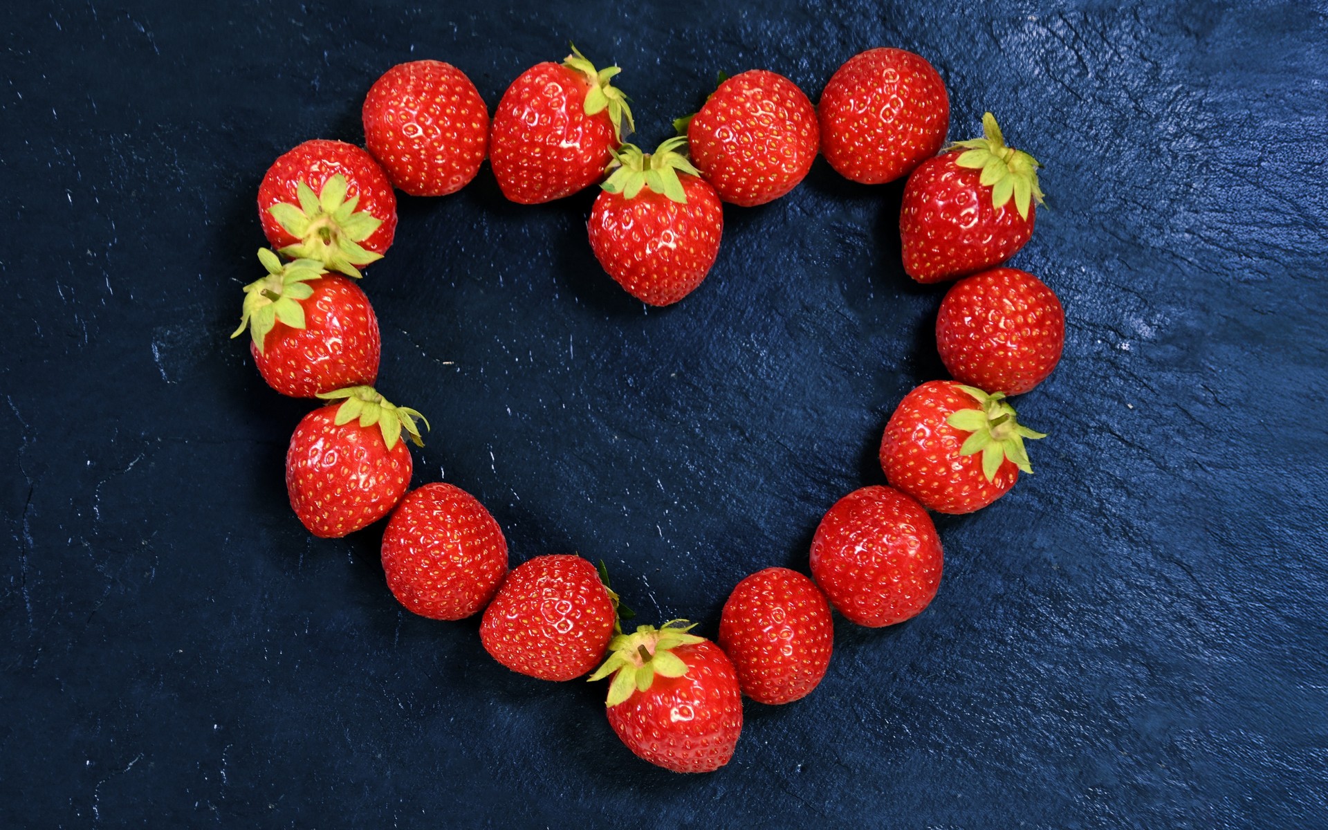 Ein Herz aus frischen Erdbeeren liegt auf einem Schieferhintergrund, der einen sehr dunklen Blau-Schwarzton zeigt.